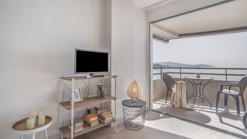 A modern living room with a flat-screen TV, books, a small table with candles, and a view of the sea through a balcony.