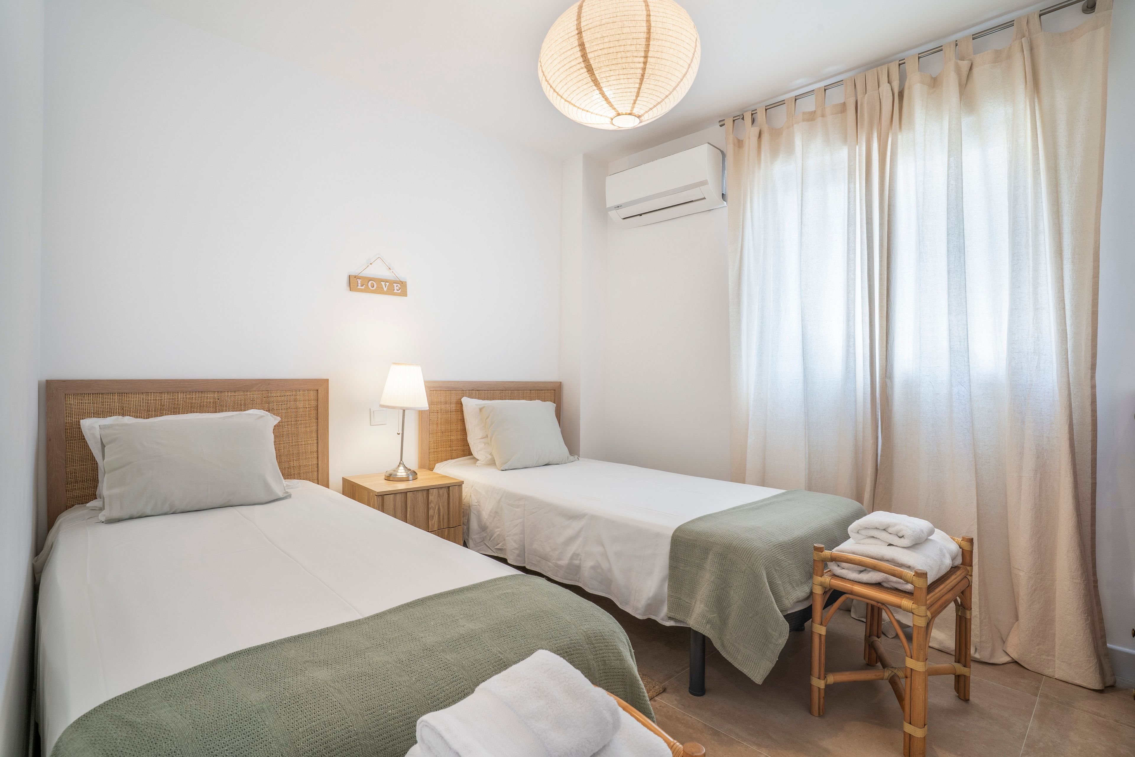 A room with two single beds, a bedside table with a lamp, a 'LOVE' sign, an air conditioning unit, and a window with curtains.