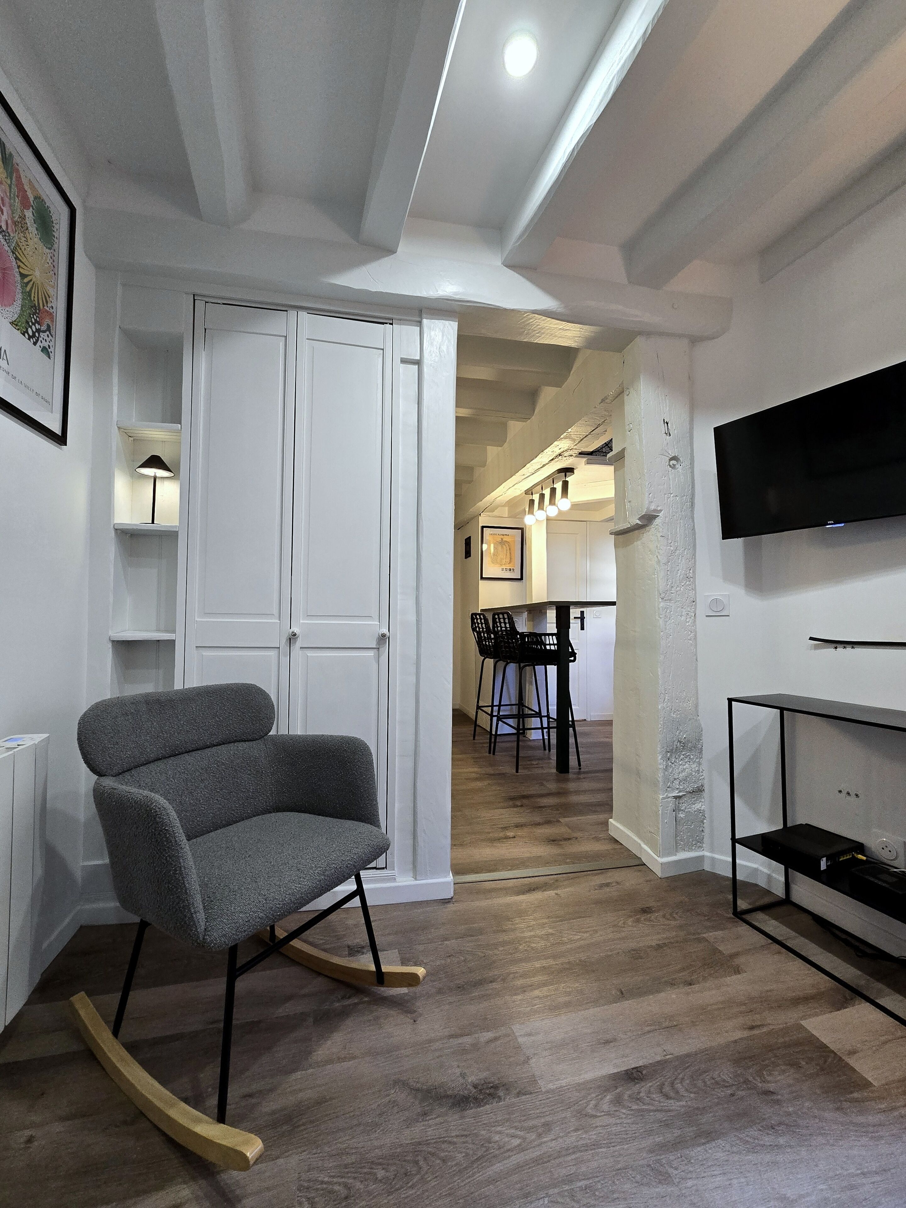 A modern living room with a grey rocking chair, a flat-screen TV mounted on the wall, and a wooden floor.
