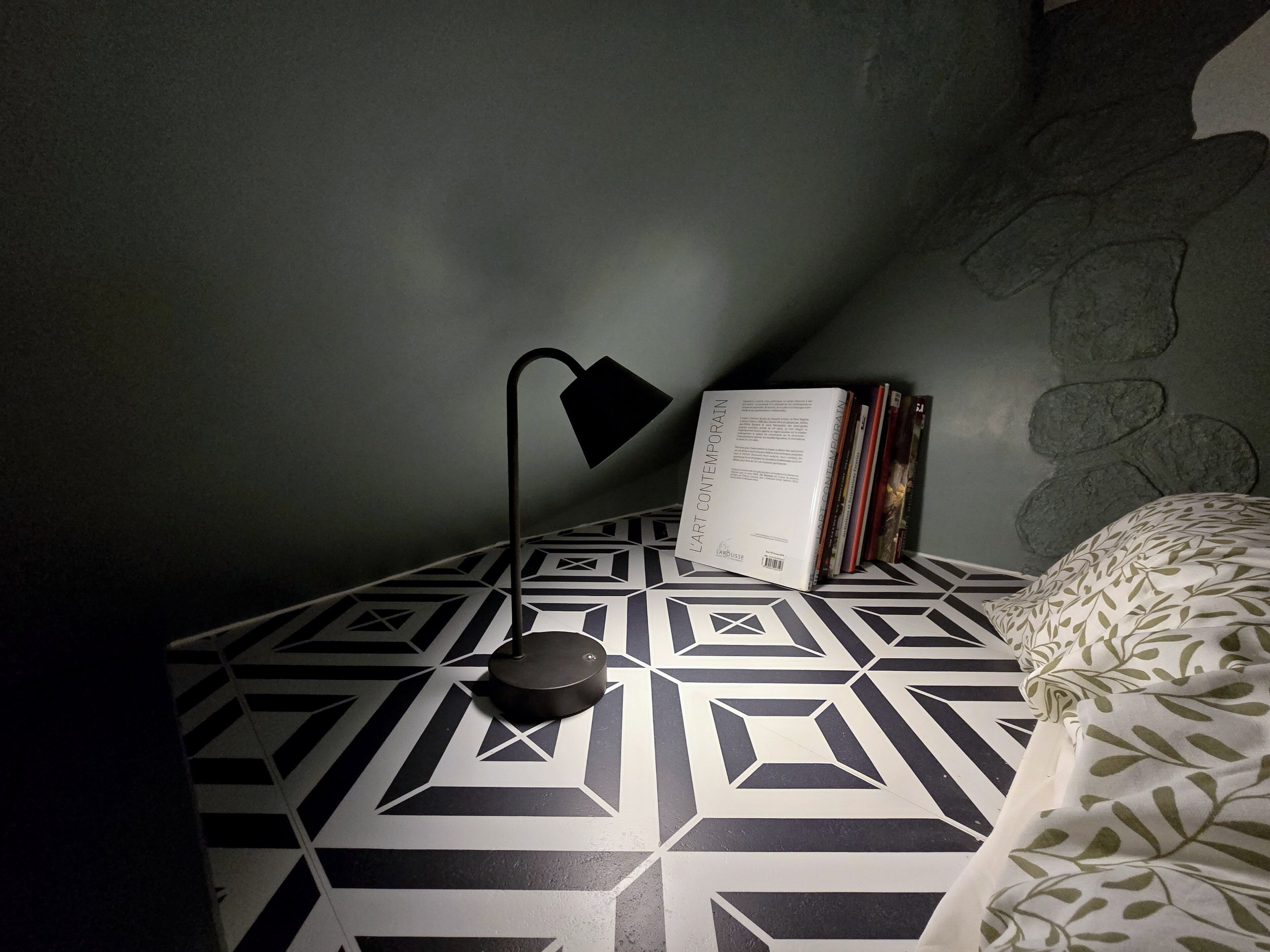 Une pièce avec un sol à motifs, une lampe de chevet, une pile de livres et un oreiller orné d’un motif de feuilles.