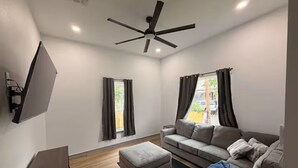 A modern living room with a sofa, ottoman, ceiling fan, and a flat-screen TV mounted on the wall. - Modern Stay Next to Downtown  Sleeps 7 (San Antonio)