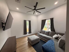 A modern living room with a sofa, ottoman, ceiling fan, and a flat-screen TV mounted on the wall. - Modern Stay Next to Downtown  Sleeps 7 (San Antonio)