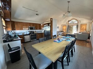 A modern kitchen with wooden cabinets, stainless steel appliances, and a dining area with a wooden table and chairs. - Surfivin': Central location• Beach Club Access • Dining • Golf (Nags Head)