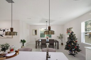 Un espace repas comprenant une table, des chaises, des luminaires suspendus, un sapin de Noël et une fenêtre avec des stores.