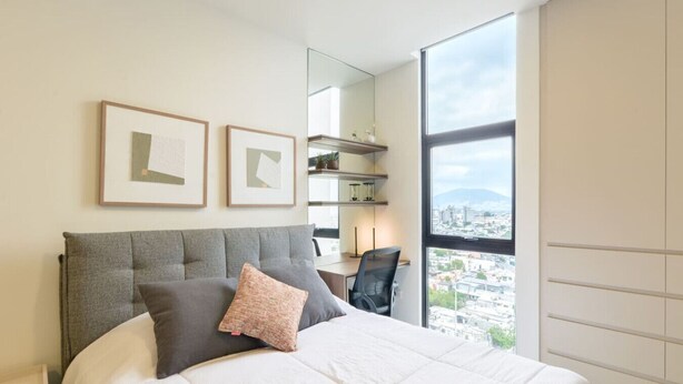 Ein modernes Schlafzimmer mit einem großen Bett, einem Schreibtisch und Blick auf eine Stadtkulisse durch ein großes Fenster.