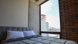 A bedroom with a large bed, a concrete wall, and a view of a cityscape through a window. - ¡Alberca y Gym! / Barrio W / 2 personas #Centro (Monterrey)