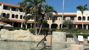 Un área de piscina con un muro de piedra, palmeras y un edificio con ventanas arqueadas al fondo.