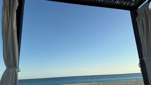 Vista de la playa desde un espacio techado con piso de arena y cielo despejado.
