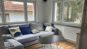Ein modernes Wohnzimmer mit einem grauen Sofa, einem runden Couchtisch und großen Fenstern.