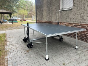 A portable ping pong table with wheels, set on a paved surface near a building with a brick wall.