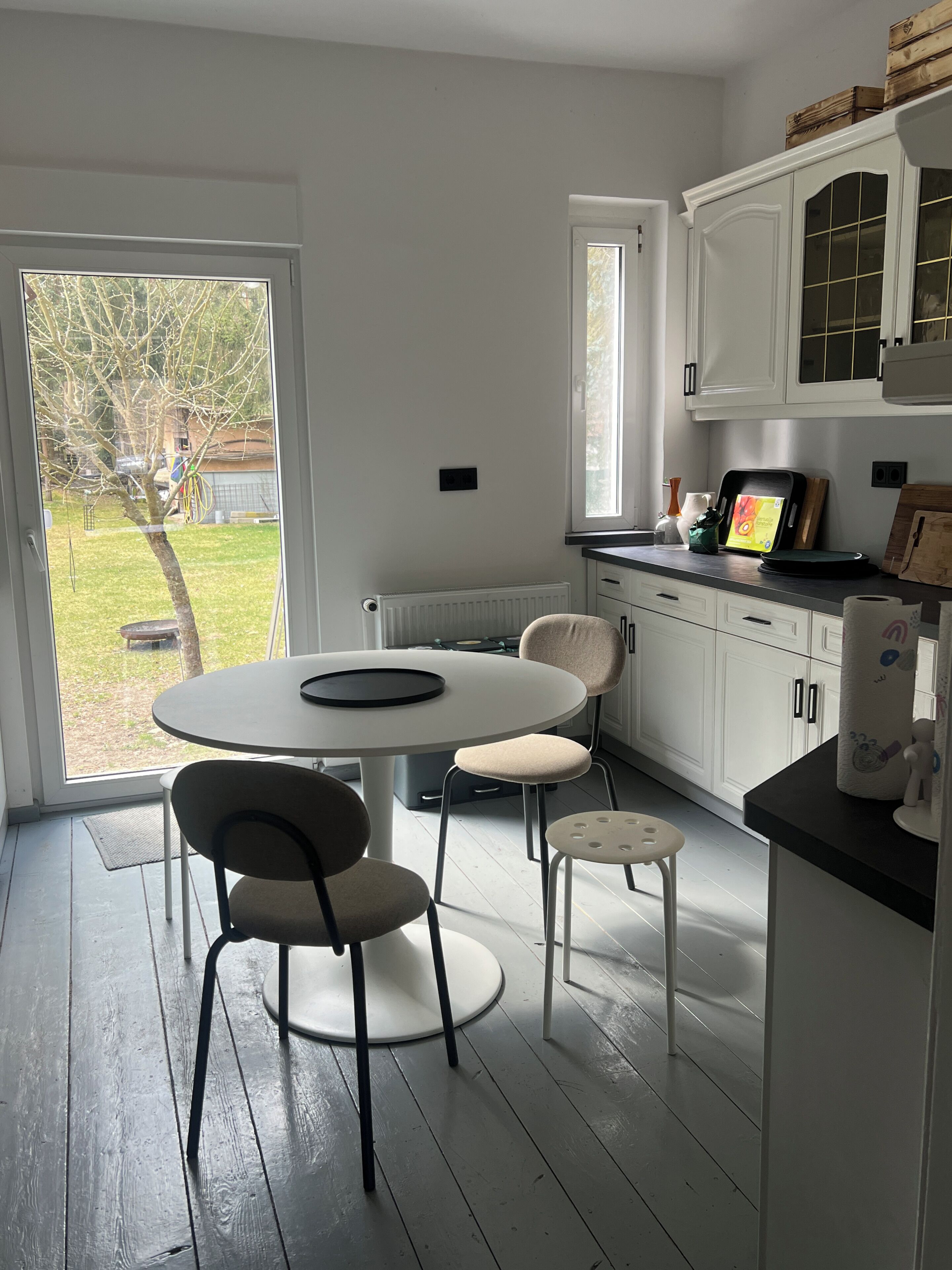 Une cuisine avec des meubles blancs, une table à manger avec des chaises, une fenêtre donnant sur l’extérieur et un plan de travail sur lequel sont disposés divers objets.