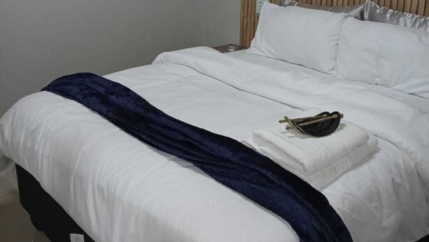 A bed with white bedding, a dark blue throw, and a pair of sunglasses on the pillow. - Pilgrims Corner Guest House  (Westonaria)