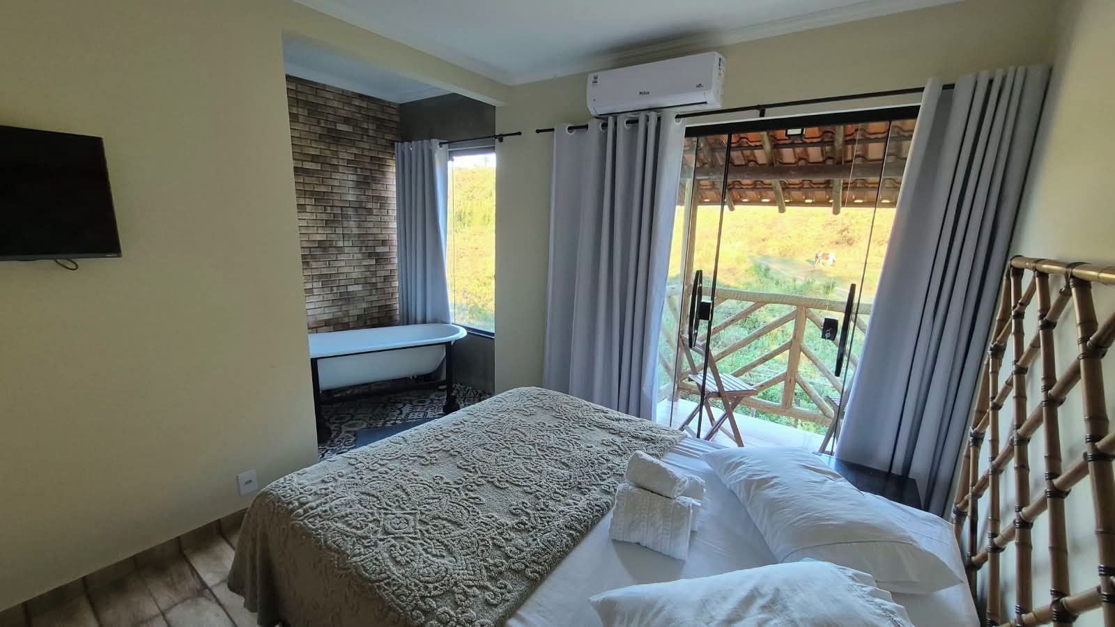 A bedroom with a bed, a small table, a window with curtains, and a view of the outdoors.