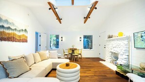 A modern living room with a sectional sofa, a round ottoman, a glass coffee table, and a fireplace. There are multiple windows providing natural light and views of the outdoors.