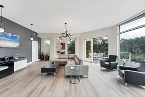 A modern living room with a sofa, coffee table, and chairs, a dining area with a table and chairs, a kitchen with a countertop and cabinets, and large windows offering a view of the outdoors. - Grand 3 Bedroom Corner suite (Orlando)