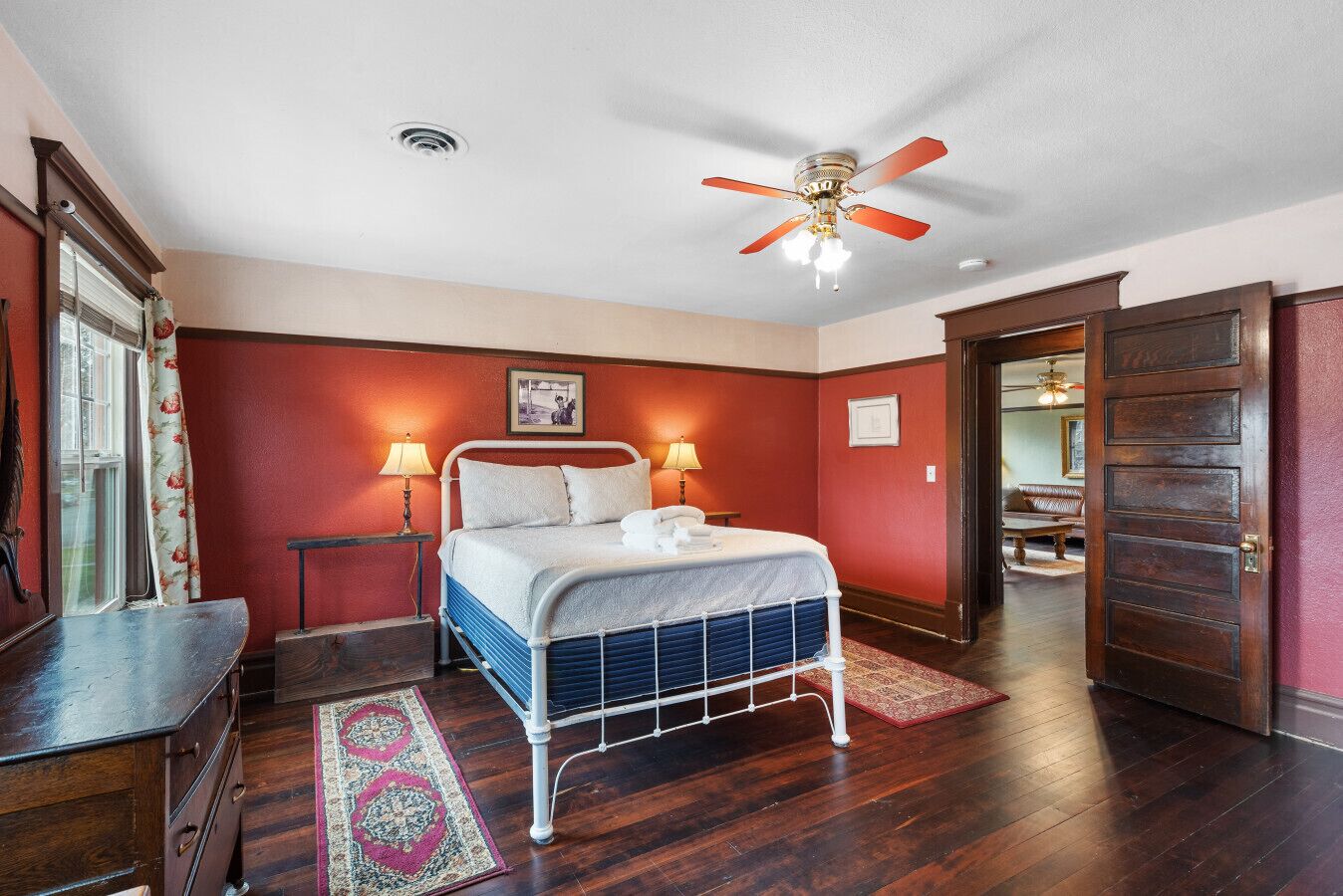 A hotel room with a bed, two bedside lamps, a ceiling fan, a wooden door, and a view into the hallway.