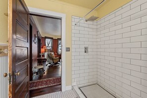 A bathroom with white subway tiles, a shower, and a visible door leading to a living area with a red carpet and furniture.