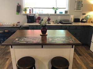 Una cocina con una isla central, una cafetera, un florero con flores y un ventanal con plantas.