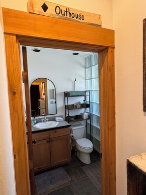 Une salle de bain avec une porte en bois portant l’inscription « Toilettes », un lavabo, un miroir, des toilettes et une étagère avec des articles de toilette.