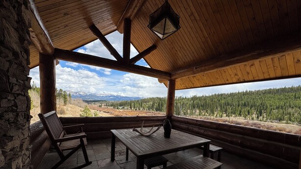 Una cabaña de madera con un muro de piedra, una mesa de madera, dos mecedoras y vistas a un bosque y montañas.