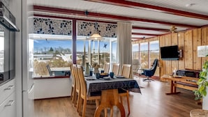 Ein Essbereich mit Holztisch und -stühlen, einem an der Wand montierten Fernseher und Blick durch große Fenster.
