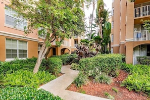 Un patio con un sendero, rodeado de abundante vegetación y un edificio de varios pisos con balcones.