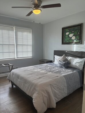 Un dormitorio con cama, mesita de noche, ventilador de techo y ventana con persianas.