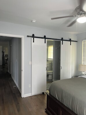 A bedroom with a bed, a ceiling fan, a window with blinds, and a bathroom visible through an open door. - Renovated House 9 Miles from Downtown ATL, Great Backyard on a Cul-de-sac! (Atlanta)
