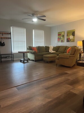 A living room with a sectional sofa, a small table, and a floor lamp. There are two windows with blinds, a ceiling fan, and wall art. - Renovated House 9 Miles from Downtown ATL, Great Backyard on a Cul-de-sac! (Atlanta)