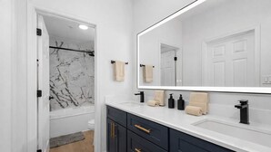 A modern bathroom with a large mirror, dark cabinets, a white countertop, and a shower with a marble wall.