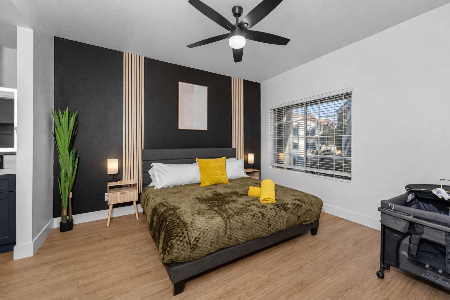Un dormitorio moderno con una cama grande, un ventilador de techo, una ventana con persianas y un escritorio pequeño con una lámpara.