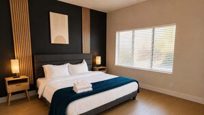 A modern bedroom with a large bed, bedside lamps, a window with blinds, and a dark headboard.