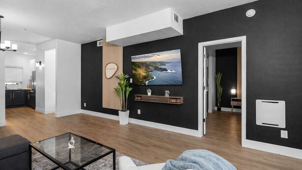 A modern living room with a large TV, a wooden accent wall, a plant, and a kitchen in the background.