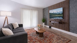 A modern living room with a sofa, a coffee table, a TV mounted on a wooden wall, and a large window with sheer curtains. - Chic South Tampa Retreat  2 King Beds  Pool, Gym & Prime Walkable Location (Tampa)