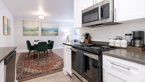 A modern kitchen with white cabinets, a black stove, a microwave, and a coffee maker. There is a dining area with a table and chairs, and a rug on the floor. - Chic South Tampa Retreat  2 King Beds  Pool, Gym & Prime Walkable Location (Tampa)