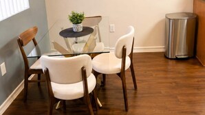 Un comedor con una mesa de vidrio, dos sillas blancas, una pequeña planta en maceta y un cubo de basura de acero inoxidable.