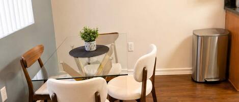 A dining area with a glass table, two white chairs, a small potted plant, and a stainless steel trash can.