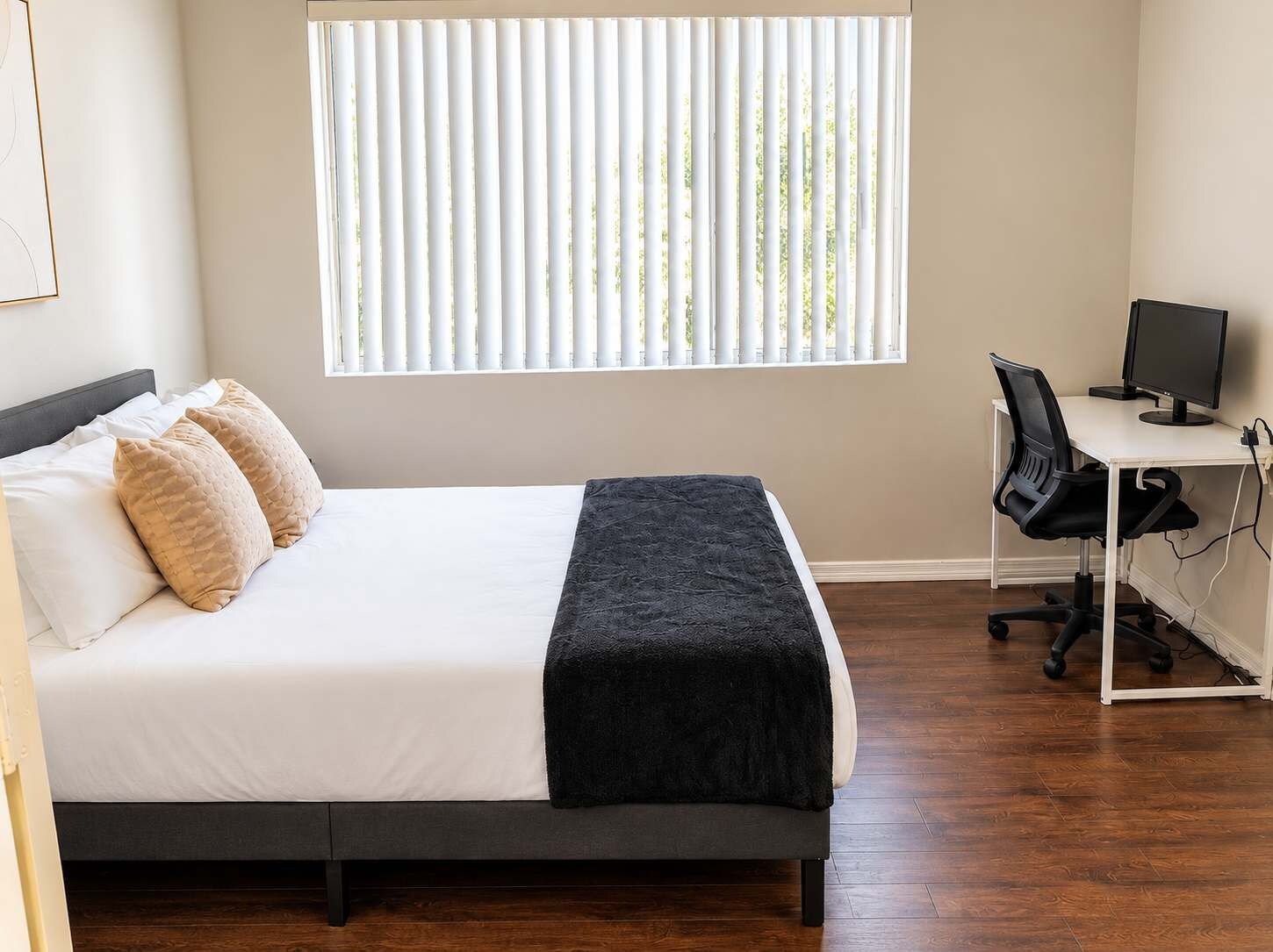 A bedroom with a bed, pillows, a desk with a computer, and a window with blinds.