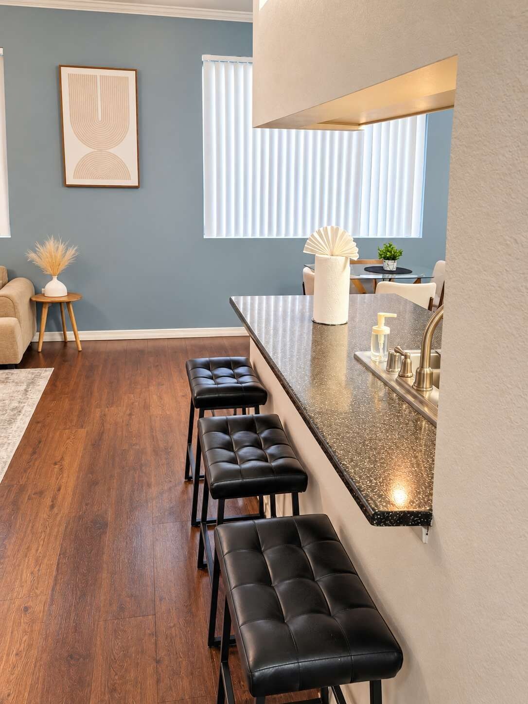 A modern kitchen with a granite countertop, a built-in oven, and a stainless steel sink. There are three black bar stools and a white vase with dried plants on a small wooden table.