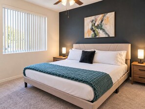 Ein Schlafzimmer mit einem Bett, zwei Nachttischen, einem Deckenventilator, einem Fenster mit Jalousien und einem an der Wand hängenden abstrakten Gemälde.