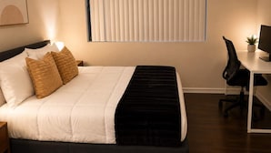 Una habitación de hotel con cama, mesita de noche, lámpara, escritorio con computadora y ventana con persianas.