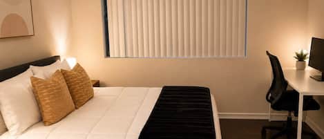 Une chambre d’hôtel comprenant un lit, une table de chevet, une lampe, un bureau avec un ordinateur et une fenêtre avec des stores.