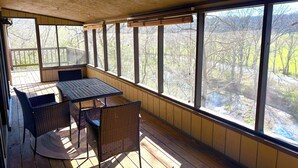 Ein überdachter Außenbereich mit Tisch und Stühlen, großen Fenstern und Blick auf Bäume sowie ein Gewässer.