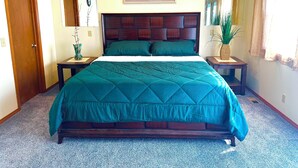 Ein Schlafzimmer mit einem großen Bett, einem Holzkopfteil, zwei Nachttischen, einem Beistelltisch mit Pflanze, einem gerahmten Bild an der Wand und einer Tür.