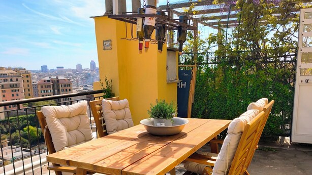 A wooden table with cushioned chairs, a potted plant, and wine bottles on a balcony with a city view. - Casa Adamo- Rooftop Hideaway (Genova)