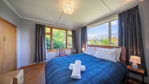 Habitación de hotel con cama, mesita de noche, lámpara, ventana con cortinas y vistas al exterior con vegetación.