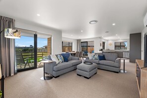 A spacious living area with a large sliding glass door to a balcony, a grey sectional sofa, a small round table, and a kitchen area with a sink and stove. - Riverside Retreat - Waipu Holiday Home (Waipu)