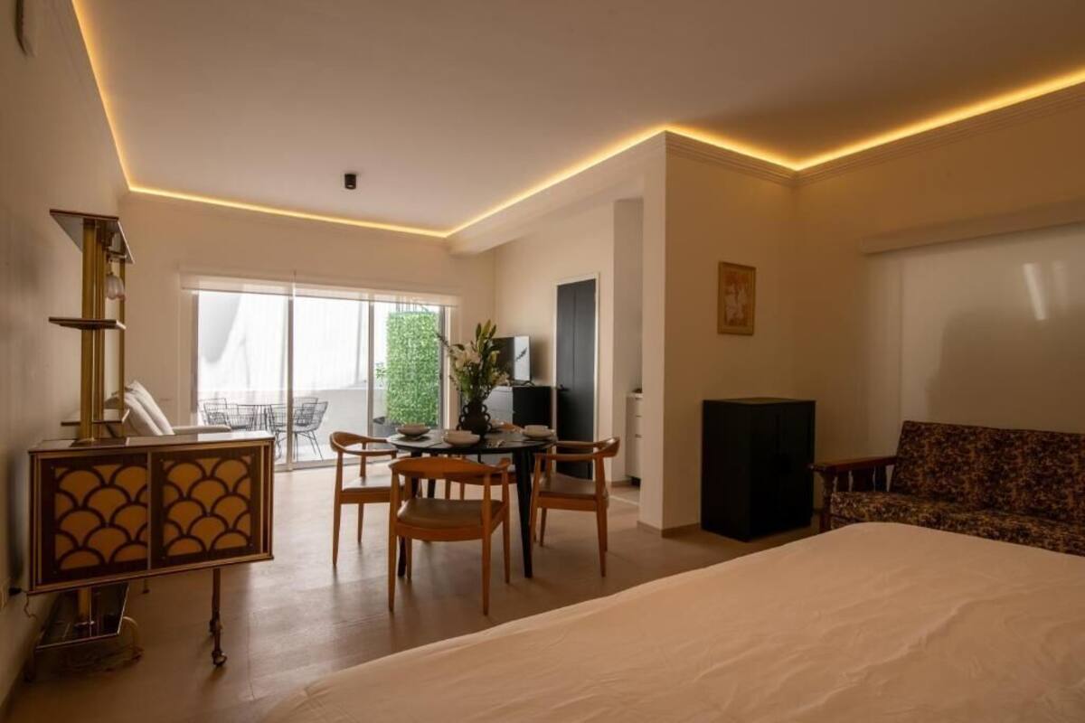 A modern hotel room with a large bed, a dining area with chairs, a TV, and a sofa.