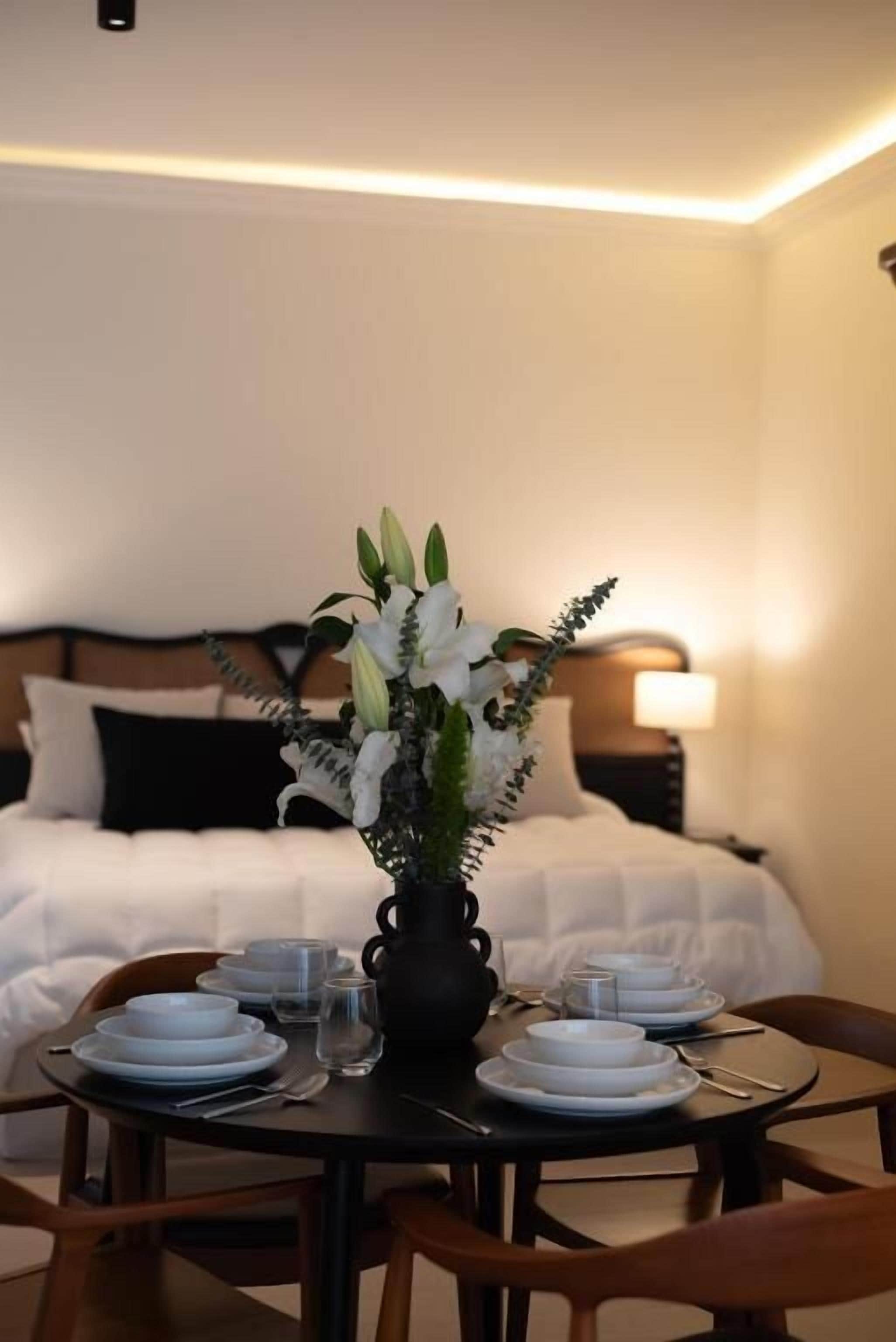 A hotel room with a neatly set dining table, a vase of flowers, and a bed with white bedding.