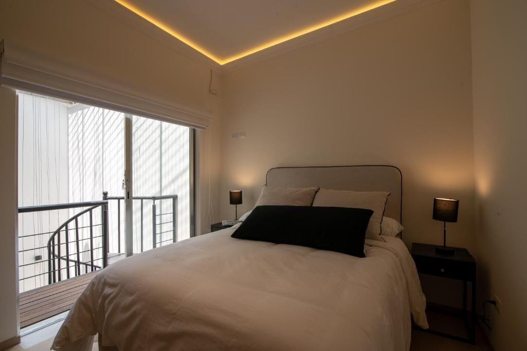 Une chambre à coucher avec un lit, deux lampes de chevet, une fenêtre donnant sur un balcon et un luminaire au plafond.